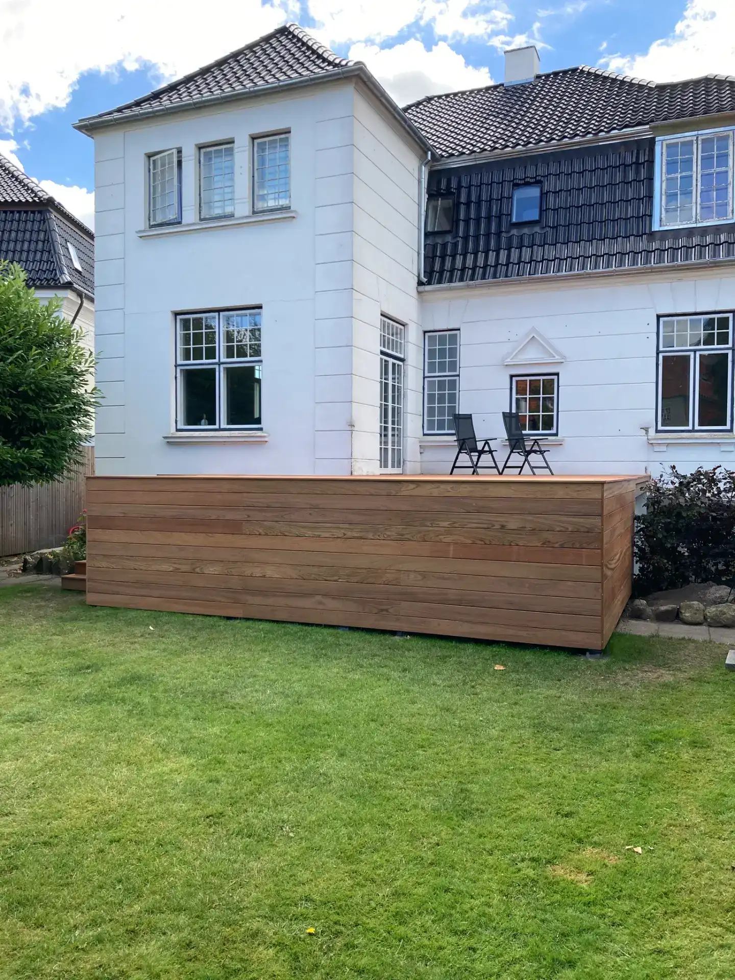 Træhegn foran hus med grøn have og udendørs siddeplads.